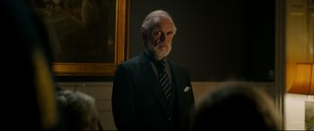 Movie still from “Journal 64” (2018), directed by Christoffer Boe – An older man wearing a suit and tie in front of a painting; Medium shot, Over the shoulder angle
