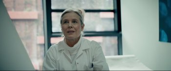 Movie still from “Journal 64” (2018), directed by Christoffer Boe – An older woman wearing a white lab coat; Close Up shot, Over the shoulder angle