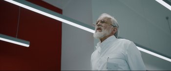 Movie still from “Journal 64” (2018), directed by Christoffer Boe – An older man with a beard and glasses is standing; Close Up shot, Low angle