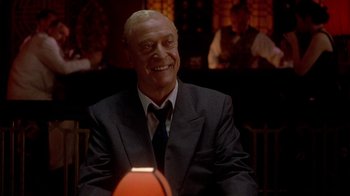 Movie still from “The Quiet American” (2002), directed by Phillip Noyce – A man in a suit and tie sitting in front of a basketball; Medium shot, Over the shoulder angle