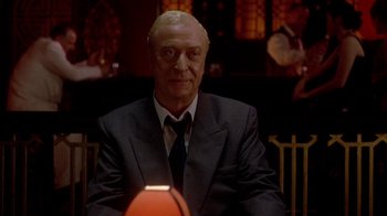 Movie still from “The Quiet American” (2002), directed by Phillip Noyce – A man in a suit and tie sitting in front of a basketball; Medium shot, Over the shoulder angle