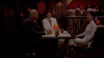 Movie still from “The Quiet American” (2002), directed by Phillip Noyce – Two men sitting at a table in front of a crowd; Medium shot, High angle