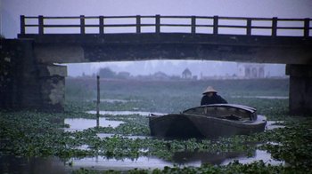Movie still from “The Quiet American” (2002), directed by Phillip Noyce – A person in a boat on a body of water near a bridge; Extreme Wide shot, High angle