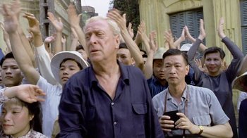Movie still from “The Quiet American” (2002), directed by Phillip Noyce – A group of people standing in front of a building with their hands up; Medium shot, Over the shoulder angle