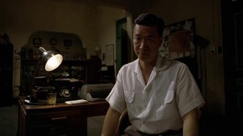 Movie still from “The Quiet American” (2002), directed by Phillip Noyce – A man sitting in front of a desk in a room; Medium shot, Over the shoulder angle