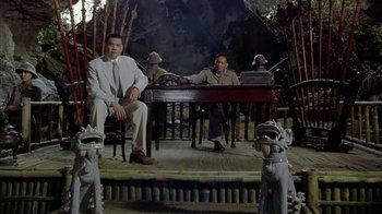Movie still from “The Quiet American” (2002), directed by Phillip Noyce – Two men sitting at a table in front of statues; Wide shot, Low angle