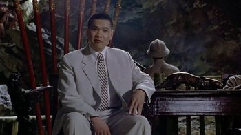 Movie still from “The Quiet American” (2002), directed by Phillip Noyce – A man in a suit and tie sitting in front of a statue; Medium shot, Low angle