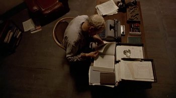 Movie still from “The Quiet American” (2002), directed by Phillip Noyce – An older man sitting at a desk with papers and a typewriter; Medium shot, Overhead angle