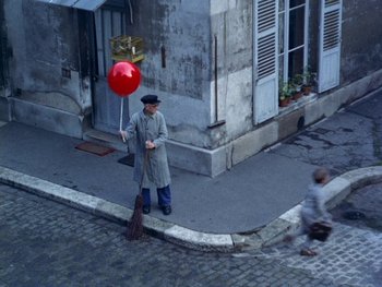 Movie still from “The Red Balloon” (1956), directed by Albert Lamorisse – A man holding a red balloon while standing on the sidewalk; Extreme Wide shot, Overhead angle