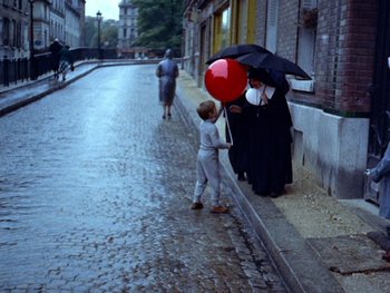 Movie still from “The Red Balloon” (1956), directed by Albert Lamorisse – Two women and a child stand on the side of the street holding an umbrella; Wide shot, High angle