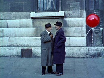 Movie still from “The Red Balloon” (1956), directed by Albert Lamorisse – Two older men standing next to each other on the sidewalk; Wide shot, High angle