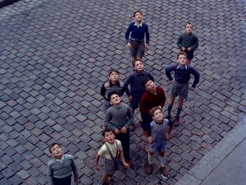 Movie still from “The Red Balloon” (1956), directed by Albert Lamorisse – A group of young men standing on a brick walkway; Wide shot, Overhead angle