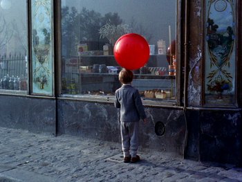 Movie still from “The Red Balloon” (1956), directed by Albert Lamorisse – A little boy standing in front of a store window holding a red balloon; Wide shot, High angle