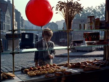 Movie still from “The Red Balloon” (1956), directed by Albert Lamorisse – A boy standing in front of a table of donuts; Wide shot, Overhead angle