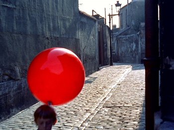 Movie still from “The Red Balloon” (1956), directed by Albert Lamorisse – A person holding a red balloon in the street; Wide shot, High angle