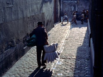 Movie still from “The Red Balloon” (1956), directed by Albert Lamorisse – A man walking down a brick street with a bunch of cardboard boxes; Wide shot, High angle