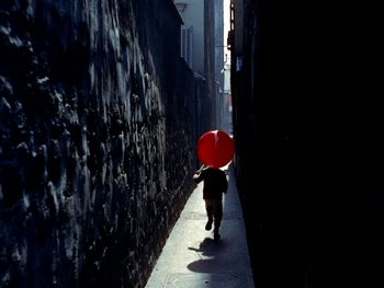 Movie still from “The Red Balloon” (1956), directed by Albert Lamorisse – A person walking down a street holding a red balloon; Extreme Wide shot, Overhead angle