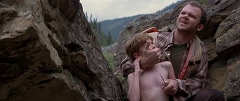 Movie still from “The River Wild” (1994), directed by Curtis Hanson – A young man is shaving his face while a man is holding a knife; Medium shot, Over the shoulder angle