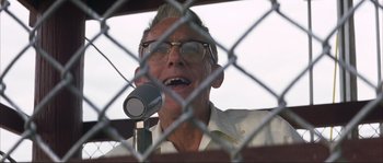Movie still from “The Rookie” (2002), directed by John Lee Hancock – An older man is speaking into a microphone in front of a chain link fence; Close Up shot, Low angle