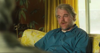 Movie still from “The Savages” (2007), directed by Tamara Jenkins – A man sitting on a couch in front of a yellow curtain; Medium shot, Over the shoulder angle