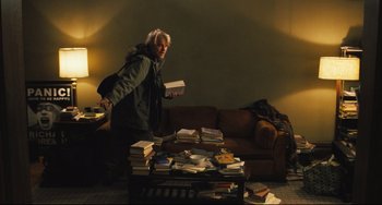 Movie still from “The Savages” (2007), directed by Tamara Jenkins – A person standing in front of a pile of books on a coffee table; Wide shot, High angle