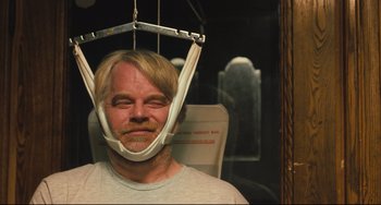 Movie still from “The Savages” (2007), directed by Tamara Jenkins – A man wearing a head hammock while sitting in a chair; Close Up shot, Over the shoulder angle