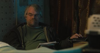 Movie still from “The Savages” (2007), directed by Tamara Jenkins – A man sitting on a couch holding a book; Medium shot, Over the shoulder angle