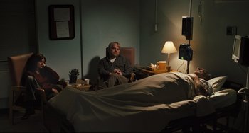 Movie still from “The Savages” (2007), directed by Tamara Jenkins – A man sitting in a chair in a room with an empty bed; Wide shot, High angle