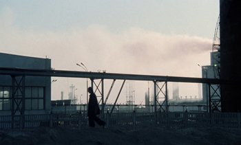 Movie still from “The Scar” (1976), directed by Krzysztof Kieslowski – A man walking across a bridge in front of an oil refinery; Extreme Wide shot, Low angle