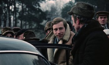 Movie still from “The Scar” (1976), directed by Krzysztof Kieslowski – A group of men standing next to each other near a car; Medium shot, Over the shoulder angle