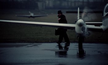 Movie still from “The Scar” (1976), directed by Krzysztof Kieslowski – A man walking across a wet runway next to an airplane; Wide shot, Low angle