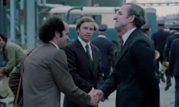 Movie still from “The Scar” (1976), directed by Krzysztof Kieslowski – A group of men in suits shaking hands outside; Medium shot, Over the shoulder angle