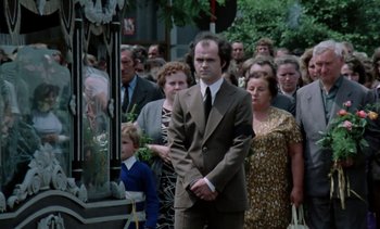 Movie still from “The Scar” (1976), directed by Krzysztof Kieslowski – A man in a suit and tie standing in front of a group of people; Wide shot, Low angle