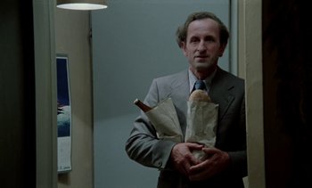 Movie still from “The Scar” (1976), directed by Krzysztof Kieslowski – A man in a suit holding a sandwich and a bottle of wine; Medium shot, Low angle