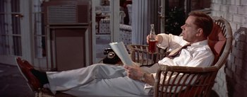 Movie still from “The Seven Year Itch” (1955), directed by Billy Wilder – An older man reading a book while holding a bottle; Medium shot, High angle