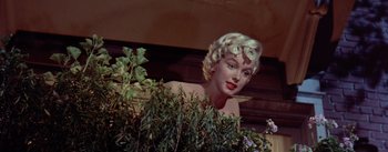 Movie still from “The Seven Year Itch” (1955), directed by Billy Wilder – A woman with blonde curly hair is standing behind a bush; Medium shot, Low angle