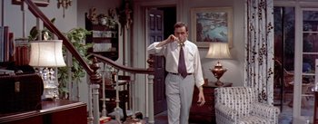 Movie still from “The Seven Year Itch” (1955), directed by Billy Wilder – A man standing in front of a staircase holding a drink; Medium shot, High angle