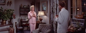 Movie still from “The Seven Year Itch” (1955), directed by Billy Wilder – A woman in a pink suit is talking to a man in a white suit in a living room; Wide shot, High angle