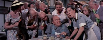 Movie still from “The Seven Year Itch” (1955), directed by Billy Wilder – An old man sitting at a table surrounded by people; Medium shot, High angle