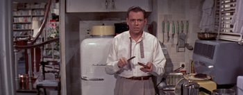 Movie still from “The Seven Year Itch” (1955), directed by Billy Wilder – A man holding a knife in a kitchen; Medium shot, Over the shoulder angle