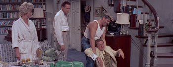 Movie still from “The Seven Year Itch” (1955), directed by Billy Wilder – Three men are in a living room and one man is sitting on a chair; Medium shot, High angle