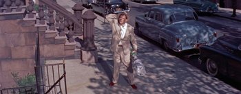 Movie still from “The Seven Year Itch” (1955), directed by Billy Wilder – A man in a tan suit and hat holding a bag on the side of the street; Wide shot, Low angle