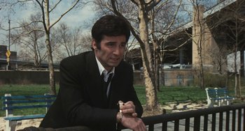 Movie still from “The Seven-Ups” (1973), directed by Philip D'Antoni – A man in a suit and tie sitting on top of a railing next to a tree; Medium shot, Over the shoulder angle