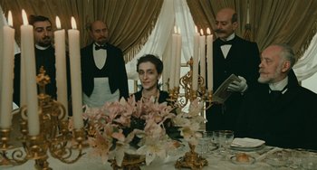 Movie still from “The Ship Sails On” (1983), directed by Federico Fellini – A woman sitting at a dinner table with two men standing next to her; Medium shot, Over the shoulder angle