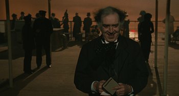 Movie still from “The Ship Sails On” (1983), directed by Federico Fellini – A man dressed as a mad hatter holding a book in his hands; Medium shot, Over the shoulder angle