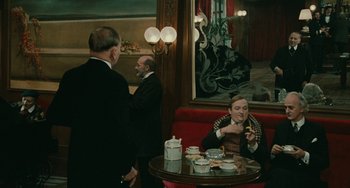 Movie still from “The Ship Sails On” (1983), directed by Federico Fellini – A man sitting at a table in front of two other men; Wide shot, Over the shoulder angle