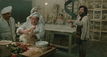 Movie still from “The Ship Sails On” (1983), directed by Federico Fellini – A group of people standing around a table with food on top of it; Wide shot, Over the shoulder angle
