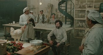 Movie still from “The Ship Sails On” (1983), directed by Federico Fellini – A group of people sitting around a table with food on top of it; Wide shot, Over the shoulder angle