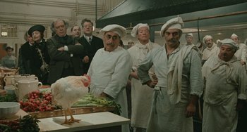 Movie still from “The Ship Sails On” (1983), directed by Federico Fellini – A group of men standing around a table with a chicken on top of it; Medium shot, High angle
