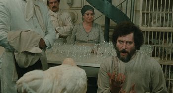 Movie still from “The Ship Sails On” (1983), directed by Federico Fellini – A group of people sitting around a table with wine glasses on top of it; Medium shot, Over the shoulder angle